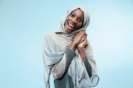 The beautiful young black african muslim girl wearing gray hijab at blue studio. She standing with mobile phone with a happy smile on her face. The human emotions, facial expression concept. Trendy colorsの写真素材