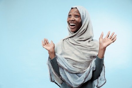 The beautiful young black african muslim girl wearing gray hijab at blue studio. She standing with a happy smile on her face. The human emotions, facial expression concept. Trendy colorsの写真素材