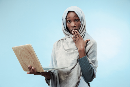Portrait Of Female University Student Working on laptop. The beautiful young black african muslim girl wearing gray hijab at blue studio. She standing with surprised emotion on her face.の写真素材