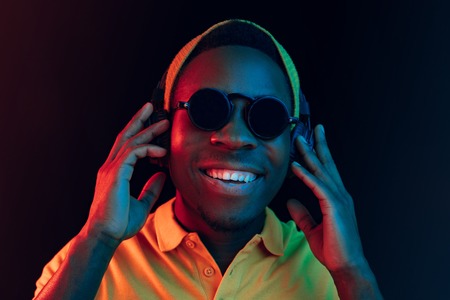 The young handsome happy hipster man listening music with headphones at black studio with neon lights. Disco, night club, hip hop style, positive emotions, face expression, dancing conceptの写真素材