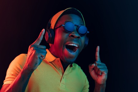 The young handsome happy hipster man listening music with headphones at black studio with neon lights. Disco, night club, hip hop style, positive emotions, face expression, dancing conceptの写真素材