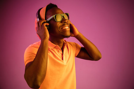 The young handsome happy hipster man listening music with headphones at black studio with neon lights. Disco, night club, hip hop style, positive emotions, face expression, dancing conceptの写真素材