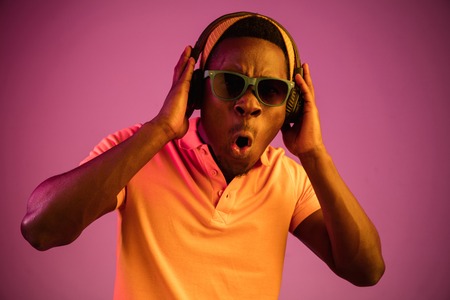 The young handsome happy hipster man listening music with headphones at black studio with neon...