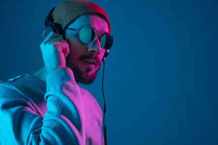 Enjoying his favorite music. Happy young stylish man in hat and sunglasses with headphones listening and smiling while standing against blue neon backgroundの写真素材