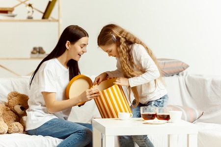 Little girl and her attractive young mother sitting on sofa with gift and spending time together at home. Generation of women. International Womens Day. Happy Mothers Day.の写真素材