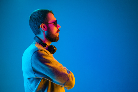 Enjoying his favorite music. Serious young stylish man in sunglasses with headphones listening sound while standing against blue neon backgroundの写真素材