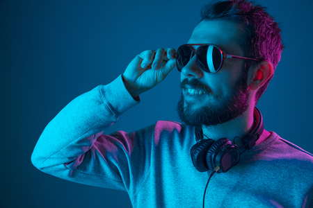 Enjoying his favorite music. Happy young stylish man in sunglasses with headphones listening and smiling while standing against blue neon backgroundの写真素材