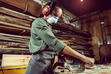 Busy and serious craftswoman grinding timbers with special machine. Beautiful woman wearing safety glasses. Concept of joiners shop and woodworking. Gender equality. Male professionの写真素材