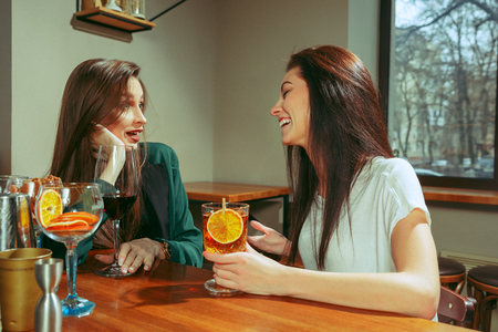 Female friends having a drinks at bar. They are sitting at a wooden table with cocktails. They are wearing casual clothes.の写真素材