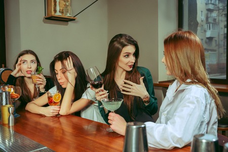 Female friends having a drinks at bar. They are sitting at a wooden table with cocktails. They are wearing casual clothes.の写真素材