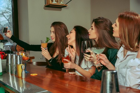 Female friends having a drinks at bar. They are sitting at a wooden table with cocktails. They are wearing casual clothes.の写真素材
