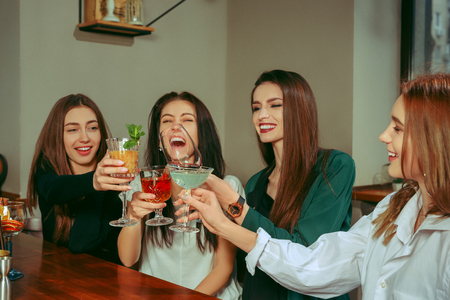Female friends having a drinks at bar. They are sitting at a wooden table with cocktails. They are clinking glassesの写真素材