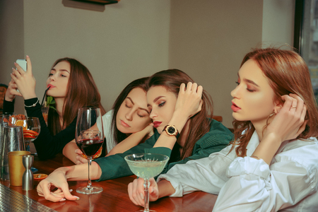 Female sad and tired friends having a drinks at bar. They are sitting at a wooden table with cocktails. They are wearing casual clothes.の写真素材