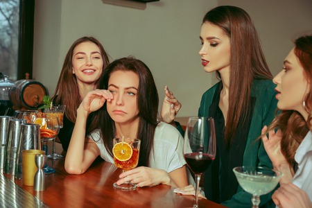 Stress. Female friends having a drinks at bar. They are sitting at a wooden table with cocktails. They are wearing casual clothes. Friends comforting and soothing a crying girl