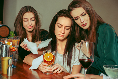Stress. Female friends having a drinks at bar. They are sitting at a wooden table with cocktails. They are wearing casual clothes. Friends comforting and soothing a crying girlの写真素材
