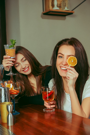 Female friends having a drinks at bar. They are sitting at a wooden table with cocktails. They are clinking glassesの写真素材