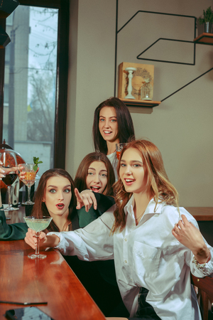Female friends having a drinks at bar. They are sitting at a wooden table with cocktails. They are wearing casual clothes.の写真素材