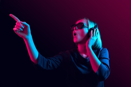 Surprised pretty woman with headphones listening to music over red neon background at studio.