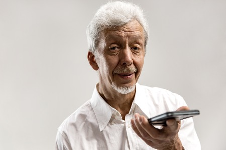 Indoor portrait of attractive senior man isolated on gray background, holding blank smartphone, using voice control, feeling happy and surprised. Human emotions, facial expression concept.の写真素材