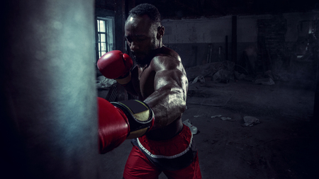 Hands of boxer over dark gym background. Strength, attack and motion concept. Fit african american model in movement. Naked muscular athlete in red gloves. Sporty man during boxingの写真素材
