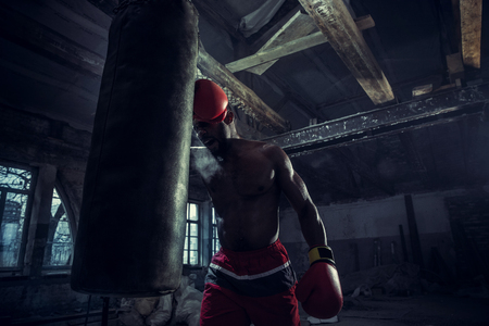 Hands of boxer over dark gym background. Strength, attack and motion concept. Fit african american model in movement. Naked muscular athlete in red gloves. Sporty man during boxingの写真素材