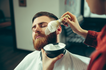 Client during beard shaving in barbershop. Female barber at salon. Gender equality. Woman in the male profession.の写真素材