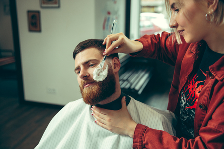 Client during beard shaving in barbershop. Female barber at salon. Gender equality. Woman in the male profession.の写真素材