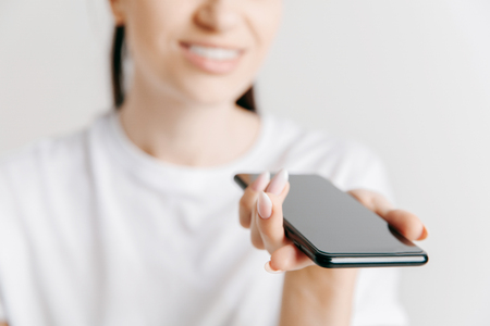 Indoor portrait of attractive young woman isolated on gray background, holding blank smartphone, using voice control, feeling happy and surprised. Human emotions, facial expression concept.の写真素材