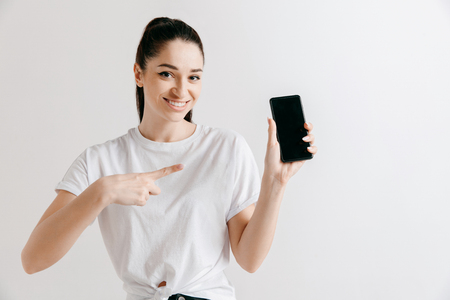 Portrait of a confident casual girl showing blank screen of mobile phone isolated over gray background at studio.の写真素材