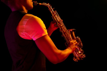 African American handsome jazz musician playing the saxophone in the studio on a neon background. Music concept. Young joyful attractive guy improvising. Close-up retro portrait.の写真素材