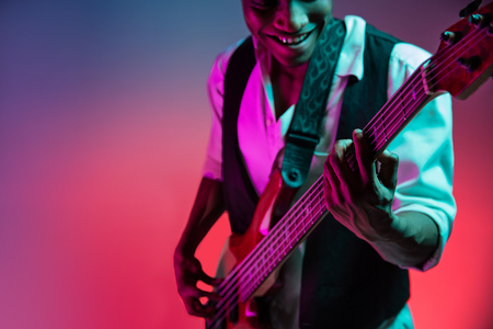 African American handsome jazz musician playing bass guitar in the studio on a neon background. Music concept. Young joyful attractive guy improvising. Close-up retro portrait.の写真素材