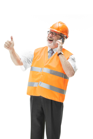 The builder in a construction vest and an orange helmet talking on a mobile phone about something. Safety specialist, engineer, industry, architecture, manager, occupation, businessman, job conceptの写真素材