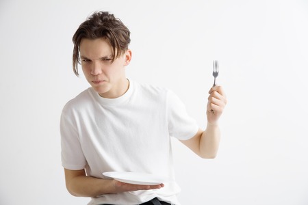 Young sad attractive caucasian guy holding empty dish and fork isolated on grey background. Copy...