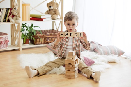 Wooden cubes with word HARD in hands of sad little boy at home. Conceptual image about education, childhood and social problems.の写真素材