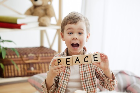 Wooden cubes with word PEACE in hands of happy smiling little boy at home. Conceptual image about...