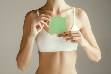 Woman health. Female model holding empty card near breast. Young adult girl with paper for sign or symbol isolated on gray studio background. Cut out part of body. Medical problem and solution.の写真素材