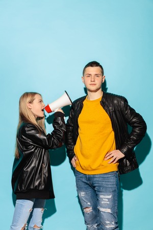 Portrait of young female student in denim clothes holding megaphone and screaming against blue studio background. Two teen friends - man and girl with serious emotion. Education in high school concept. Copy spaceの写真素材