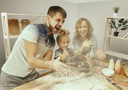 Cute little girl and her beautiful parents preparing the dough for the cake in kitchen at home....