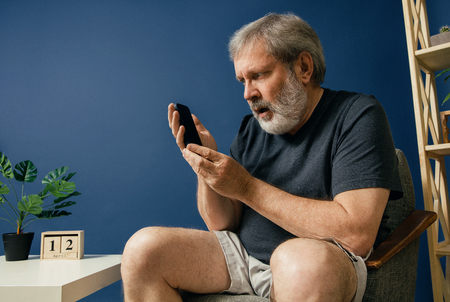 Wondering of new. Old bearded man with alzheimer desease sitting on the chair and trying to use smartphone on blue background. Concept of illness, memory loss due to dementia, healthcare, disorder.の写真素材