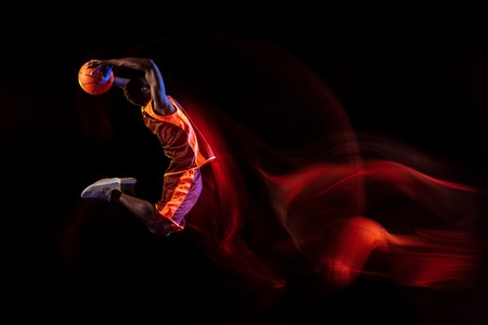 Heating the atmosphere. African-american young basketball player of red team in action and neon lights over dark studio background. Concept of sport, movement, energy and dynamic, healthy lifestyle.の写真素材