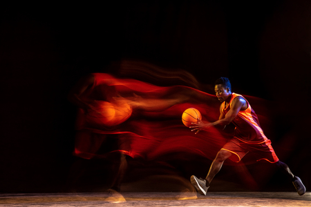 Melting track of fire. African-american young basketball player of red team in action and neon lights over dark studio background. Concept of sport, movement, energy and dynamic, healthy lifestyle.の写真素材