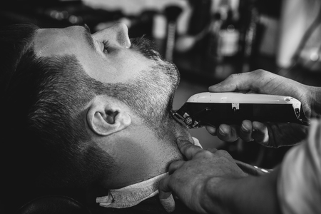 Client during beard shaving in barbershop. Young handsome barber and attractive bearded man. Black and white or colorless photo. Hairstyle, salon, hairdresser, barber shop, lifestyle concept. Caucasian male models.の写真素材