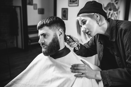 Young woman making haircut for bearded man at barbershop. Female barber at salon. Gender equality. Woman in the male profession. Black and white or colorless photo. Hairstyle, salon, hairdresser, lifestyle concept.の写真素材
