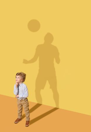 To defend or attack. Hard trainings. Childhood and dream concept. Conceptual image with child and shadow on the yellow studio wall. Little boy want to become football player and to buil a sport career.の写真素材