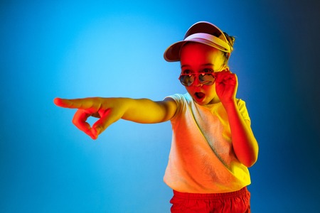 Wow. Beautiful female half-length portrait pointing to left over blue lights neon studio backgroud. Young emotional surprised teen girl standing with open mouth. Human emotions, facial expression concept. Trendy colorsの写真素材