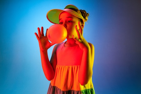 Young girl blowing bubble gum. Happy teen girl standing over trendy blue neon studio background. Beautiful female portrait. Young satisfy girl. Human emotions, facial expression, summer holidays concept.の写真素材