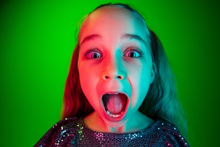 Wow. Beautiful female half-length portrait over green lights neon studio backgroud. Young emotional surprised teen girl standing with open mouth. Human emotions, facial expression concept. Trendy colorsの写真素材