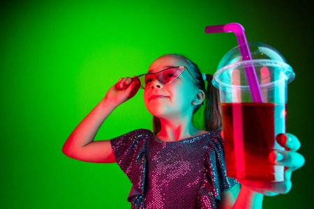 Happy teen girl standing and smiling with cola isolated on trendy green lights neon studio background. Beautiful female portrait. Young satisfy girl. Human emotions, facial expression, summer holidays concept.の写真素材