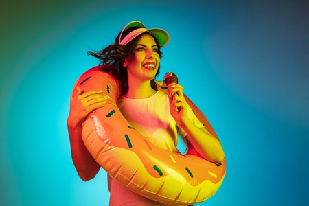 Happy young woman in a cap and in a rubber ring with an icecream on trendy blue neon studio...