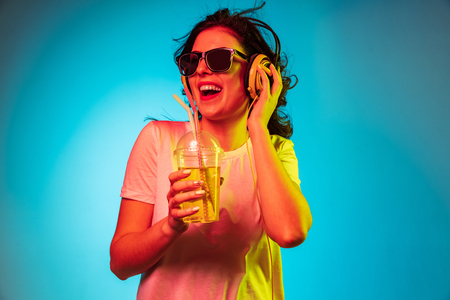 Happy young woman dancing and smiling in headphones over trendy blue neon studio background. Beautiful female portrait. Concept of human emotions, facial expression, summer holidays or weekend.の写真素材